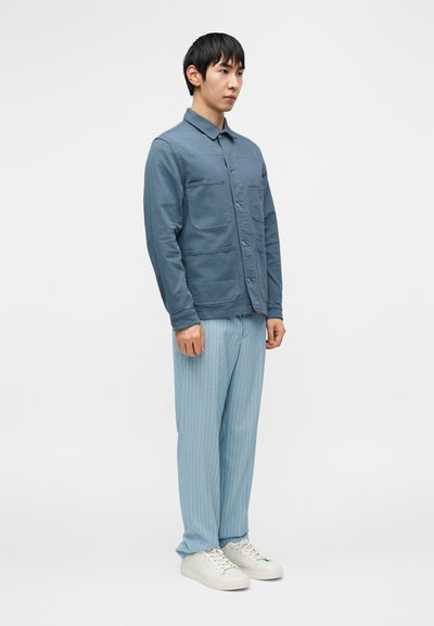 Ανοιχτό μπλε μπουφάν με κουμπιά και δύο τσέπες στο στήθος, συνδυασμένο με ανοιχτό μπλε παντελόνι με ρίγες και λευκά αθλητικά παπούτσια. Σαφής εφαρμογή και λεία υφή.