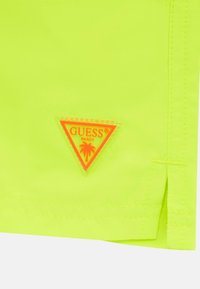 Pantaloni corti giallo neon con una patch triangolare arancione che presenta la scritta "GUESS BEACH" e un motivo di palma. Tessuto liscio e orlo cucito.
