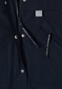 Dettaglio della giacca blu navy scuro con zip frontale, bottoni automatici, due tasche frontali con linguette di tiraggio nere con marchio "Cecil Outerwear".