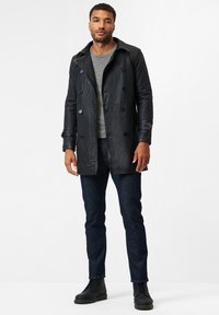 Zwarte leren dubbelzijdige trenchcoat met een licht glanzende afwerking, voorzien van knoopaccenten, gedragen over een grijze ronde hals trui en donkere jeans.