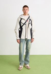 Lett, kremfarget hettejakke med svarte detaljer, glidelåslommer, brukt over en lys skjorte, kombinert med falmede blå jeans og brune joggesko.