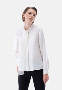 Blusa bianca a maniche lunghe con colletto alto e cravatta coordinata. Tessuto morbido con una qualità leggermente trasparente e maniche con polsini.