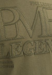 Olivgrön tyg med upphöjd, broderad text "PME LEGEND" och subtil struktur. Ytterligare text "Freedom Unlimited" är också broderad.