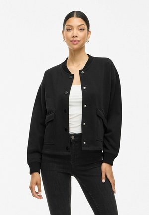 Czarny dresowy jacket z zapięciem na guziki, z prążkowanymi mankietami i dwoma przednimi kieszeniami. Gładki materiał, luźny krój i zaokrąglony dekolt.