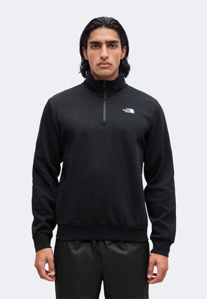 Mann mit mittellangen dunklen Haaren, der einen schwarzen Viertelzip-Pullover mit dem The North Face-Logo auf der Brust trägt und vor einem schlichten Hintergrund steht.
