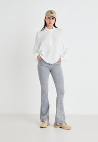 Blusa bianca, con texture e colletto ondulato, con bottoni sul davanti; abbinata a jeans grigi chiari svasati e sneakers beige; indossata con un berretto beige.
