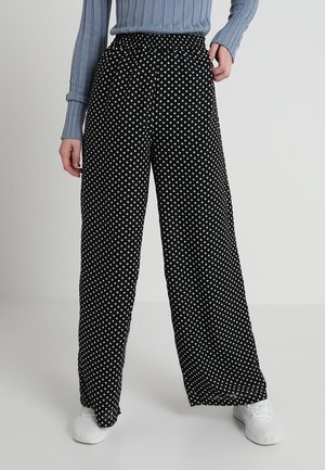 Schwarze, weit geschnittene Hosen mit weißen Punkten, kombiniert mit einem hellblauen, gerippten Pullover und weißen Sneakern, vor einem schlichten Hintergrund stehend.