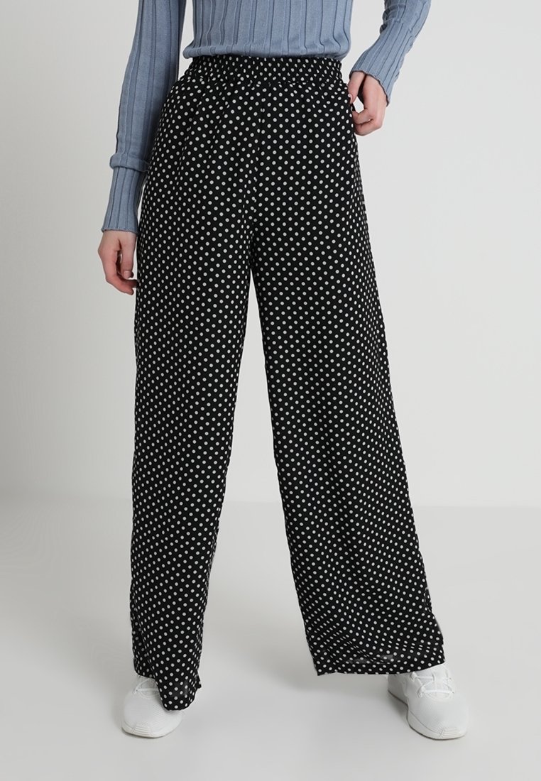 Pantalon noir à jambes larges à pois blancs, porté avec un pull côtelé bleu clair et des baskets blanches, debout devant un fond uni.