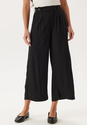Frau trägt schwarze, weit geschnittene, hoch taillierte, gekürzte Hose, kombiniert mit schwarzen Ballerinas und einem weißen ärmellosen Top.