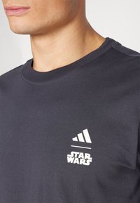 Teflon modra bombažna majica s kratkimi rokavi, ki vključuje bele logotipe Adidas in Star Wars. Ima okrogel izrez in gladko teksturo.