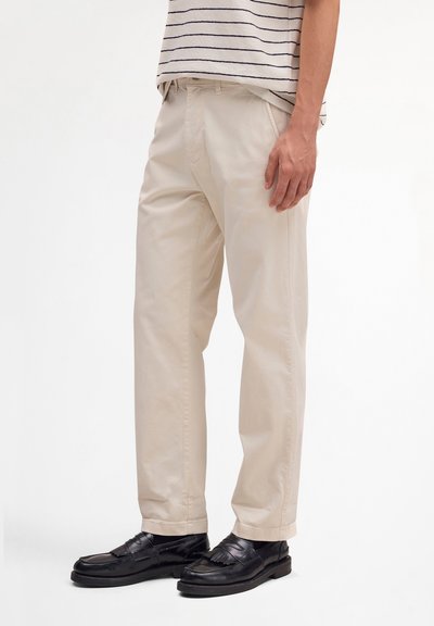Homme portant un pantalon beige à jambes droites avec des poches latérales et des mocassins en cuir noir, debout devant un arrière-plan uni.
