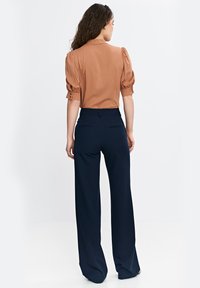 Brązowa bluzka z bufkami i akcentami guzików, połączona z granatowymi spodniami o szerokich nogawkach z tylnymi kieszeniami; gładka tkanina i dopasowany krój.