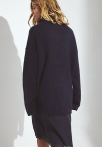 Per didelis tamsiai mėlynas megztinis su minkšta tekstūra, išsiplėtusiais rankogaliais ir apvadu. Laisvas siluetas, krentantys pečių siūlės ir minimalus detalizavimas gale.