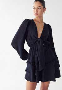 Ženska v črni teksturirani dolgi obleki z napihnjenimi rokavi, globokim V-izrezom, zavezanem v pasu in slojevitem volanastem krilu, pozira s stranjo.