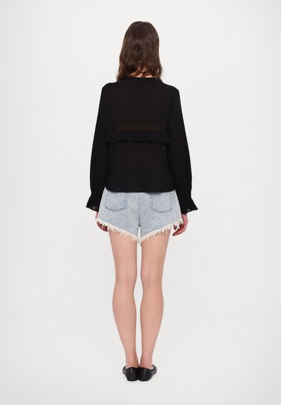 Schwarzes Langarmoberteil mit einem Rüschensaum und strukturiertem Rückenteil, kombiniert mit hellblauen, fransigen Jeansshorts. Flache schwarze Schuhe runden den Look ab.