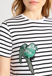 Pruhované černobílé tričko s motivem vyšívaného ptáka na přední straně, který má jemné detaily a kulatý výstřih.