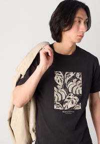 T-shirt nera in cotone con grafica di foglia di monstera color crema, con collo a giro e maniche corte, indossata da un modello con una giacca beige.