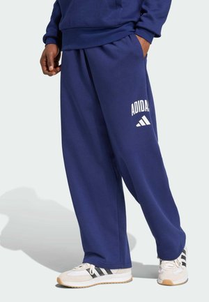 Person trägt dunkelblaue Adidas-Sweathosen und weiße Adidas-Sneaker mit schwarzen Streifen, steht mit einer Hand in der Tasche.