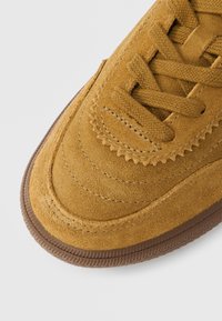 Κοντινή λήψη στο εμπρόσθιο μέρος ενός ταμπά σουέντ αθλητικού παπουτσιού με καφέ υφή σόλας από καουτσούκ και κορδόνια που ταιριάζουν, σε φωτεινό φόντο.