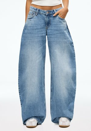 Person iført lyseblå vidde jeans og hvide sneakers, stående med hænderne i lommerne.