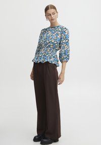 Blusa peplum floreale in blu, rosa e verde, con maniche a palloncino e vita arricciata, abbinata a pantaloni larghi marroni e scarpe nere chunky.