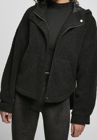 Black fleece jacket with a structured hood, front zipper, and oversized fit. Textured, soft material with side pockets and cuffed sleeves.