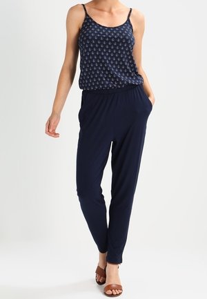 Combinaison bleu marine avec un haut sans manches à motif de petites ancres, taille élastique et jambes fuselées ; associée à des sandales marron.