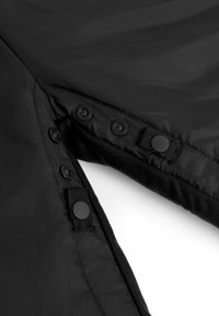 Zwarte buitenkleding van zacht nylon materiaal met drukknoopjes en een gestructureerde binnenvoering. Inclusief een veilige klittenbanddetail.