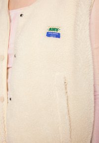 Crèmekleurige fleece vest met drukknoopjes, met een label op de borst en een textuur die lijkt op zachte, krullende wol.