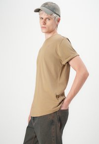 Jongere met kort blond haar draagt een beige pet, een tan t-shirt met opgerolde mouwen en donkere spijkerbroeken, poseert met hand in de zak tegen een witte achtergrond.