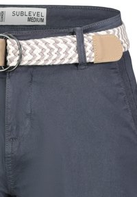 Marineblauwe broek met een gevlochten crèmekleurige elastische riem, voorzien van een ronde zilveren gesp en een tan leren accent nabij de riemlus.