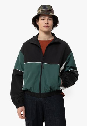 Kleurblokkering jack in zwart, groen en wit; heeft een rits, elastische manchetten en een relaxte pasvorm. G gedragen met een camouflage emmerhoed en een oranje shirt.