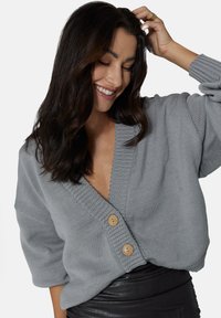 Szary dziergany kardigan z głębokim dekoltem w kształcie litery V, szerokimi rękawami i dwoma drewnianymi guzikami z przodu. Wygląda na miękki i luźny.