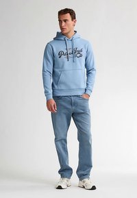 Světlé modrá mikina s kapucí, přední kapsou, šňůrkami a potiskem loga. Kombinováno se světlými džínami a bílými teniskami.