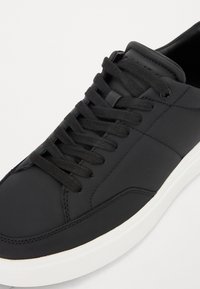 Sneaker en cuir noir avec des lacets plats noirs, une toe arrondie et une semelle en caoutchouc blanca. Présente une couture minimale et des détails latéraux subtils.