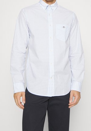 Homme portant une chemise à carreaux bleu clair et blanc à manches longues et un pantalon sombre, debout devant un fond uni.