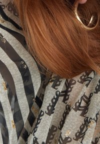 Lätt scarf i en mix av beige och svart med ränder, med ett subtilt mönster av guldfläckar och en varumärkeslogotyp.