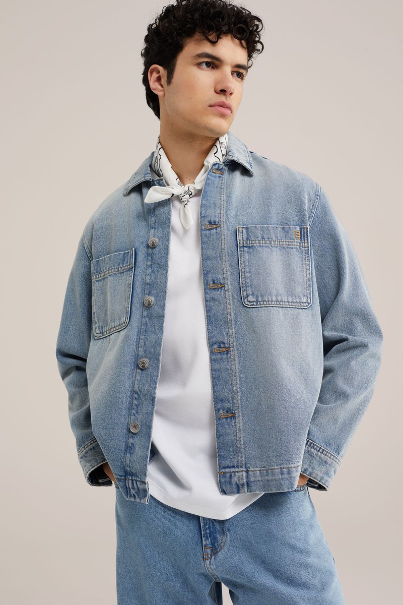 Young man wearing light blue denim jacket and jeans, white T-shirt, and white patterned neckerchief, looking away with hands in pockets.