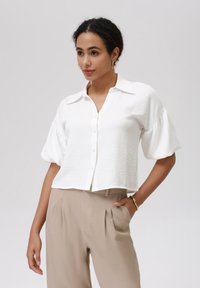 White short-sleeve shirt with textured fabric, button front, and a classic collar, paired with beige high-waisted trousers.