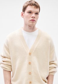 Tejszínszínű, vastag kötött kardigán V-nyakkivágással és bordázott részletekkel. Öt fa gomb az elején; bő szabás, felhajtott ujjakkal.