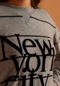 Felpa grigia con scritte nere che dicono "New York City." Presenta un collo a costine e strisce orizzontali sul tessuto.