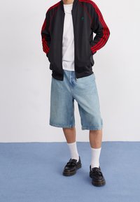 Person klädd i svart och röd Adidas-jacka, vit t-shirt, ljusblå denimshorts, vita strumpor och svarta mockaskor med tofsar stående på blått golv.