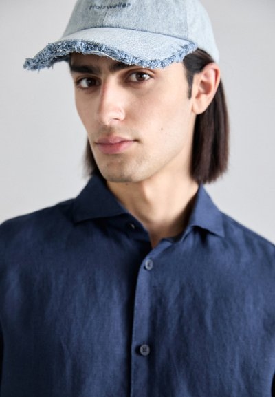 Gorra de mezclilla con borde deshilachado y logotipo, combinada con una camisa de lino azul oscuro con cuello y botones al frente. Textura suave, diseño casual.