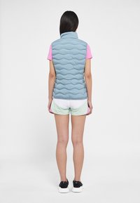 Gilet imbottito azzurro chiaro senza maniche con colletto alto, caratterizzato da dettagli trapuntati, indossato sopra una camicia rosa e pantaloncini verdi. Scarpe da ginnastica nere.