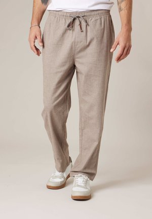 Homme portant un pantalon beige à cordon de serrage et des baskets blanches, debout les mains détendues le long du corps sur un fond neutre.