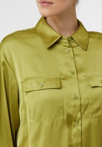 Woman wearing a shiny olive green button-up shirt with two front flap pockets and a pointed collar, close-up on the upper torso.