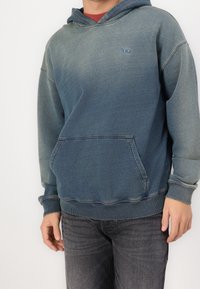 Persona con sudadera de degradado azul, con bolsillo frontal, y jeans gris oscuro, de pie contra un fondo blanco.