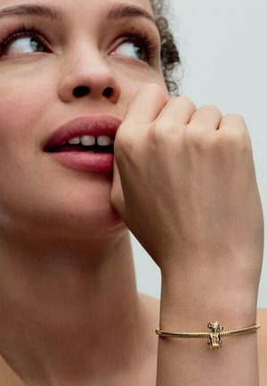 Goldenes Armband mit einem detaillierten Tieranhänger, mit einem gedrehten Design und glatter Metalloberfläche, getragen an einem Handgelenk mit heller Hautfarbe.