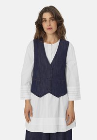 Dunkelblaue Jeansweste mit V-Ausschnitt, drei Knöpfen und zwei Fronttaschen, getragen über einem weißen Hemd mit strukturierten Ärmeln.