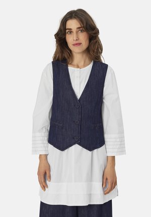 Donkerblauw denim vest met een v-hals, drie knopen en twee voorzakken, gedragen over een witte blouse met textuur mouwen.